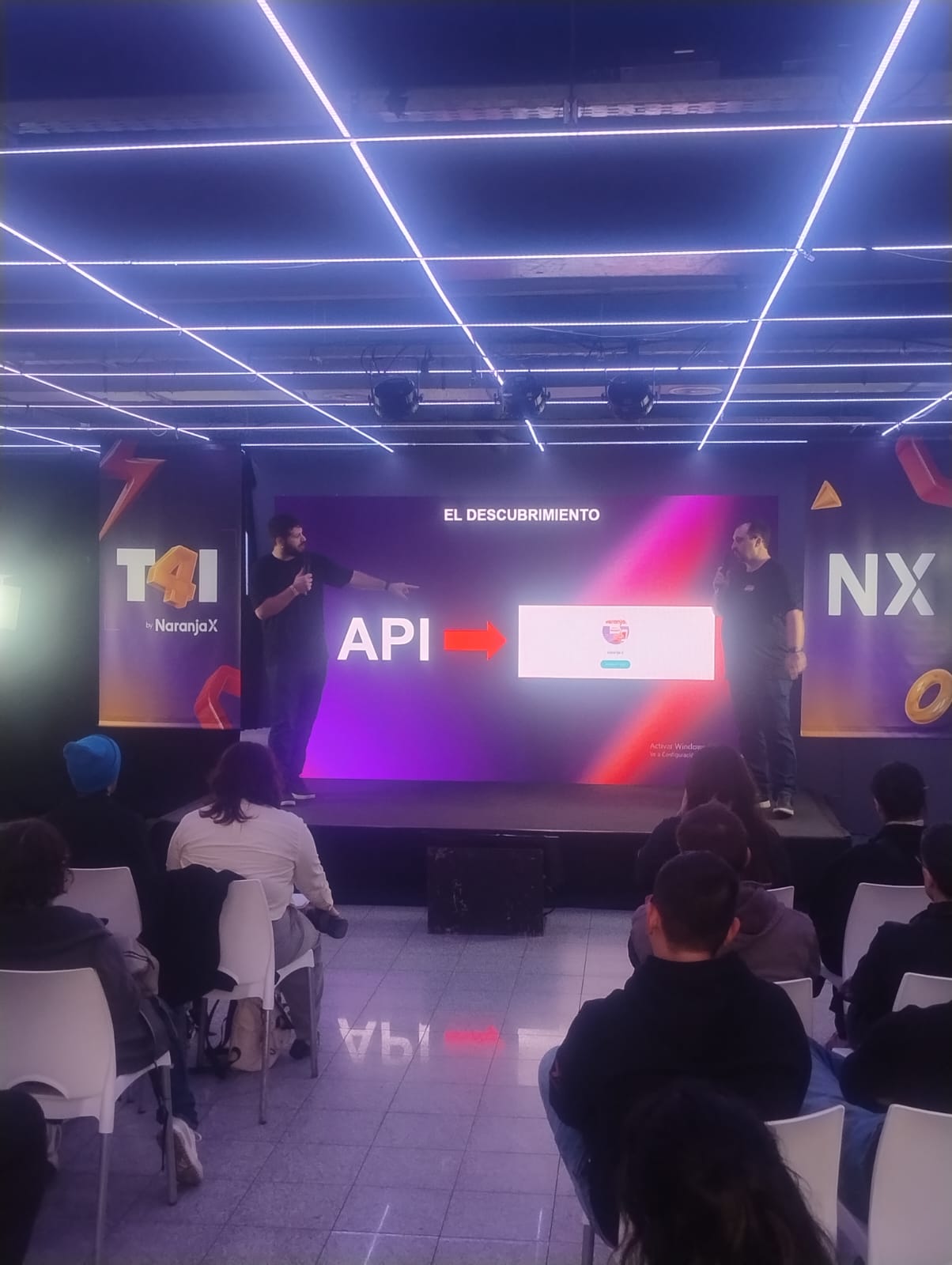 Pantalla con una presentación, dos expositores y público sentado.